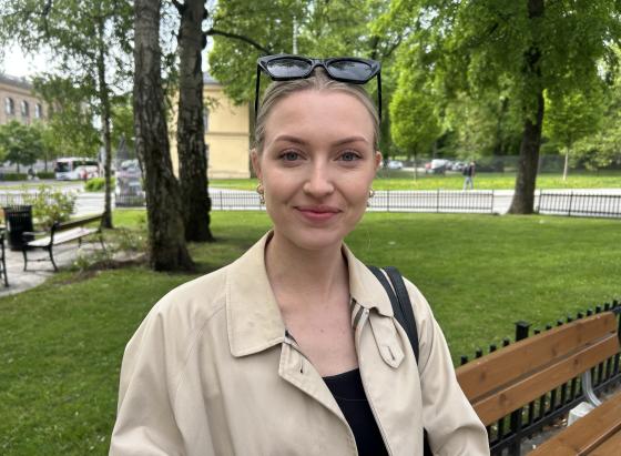 Ung dame som smiler med grønn natur i bakgrunnen imens hun venter på bussen.