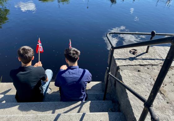To personer sitter i trapp nede ved vannet med hvert sitt norske flagg
