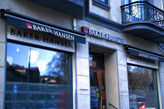 Bildet viser fasaden til en Baker Hansen-kafé med sort markise, røde «1861»-detaljer og store utstillingsvinduer som speiler trær og bygninger på andre siden av gaten. Den aktuelle Baker Hansen-kafeen på bildet er nå nedlagt.