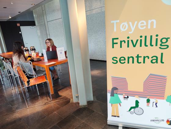 Tøyen Frivillighetssentral som plakat. Sitter to stk ved et oransje bord og prater.