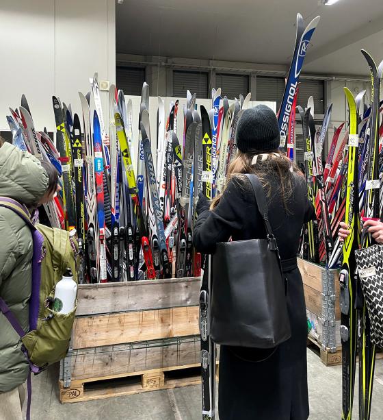 Mange langrennski stå rett opp og ned i en kasse. En kvinne står forran kassen og holder et parr med langrennsski