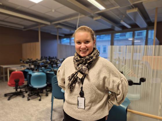 En dame står foran et rom under oppussing fylt med stoler. Hun har håret opp i en hestehale, og skjerf og genser på. Hun smiler og har hendene bak ryggen.