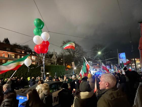 Mennesker st&aring;r langs en gate og holder flagg og ballonger i r&oslash;dt, hvit og gr&oslash;nt. Det er m&oslash;rkt ute