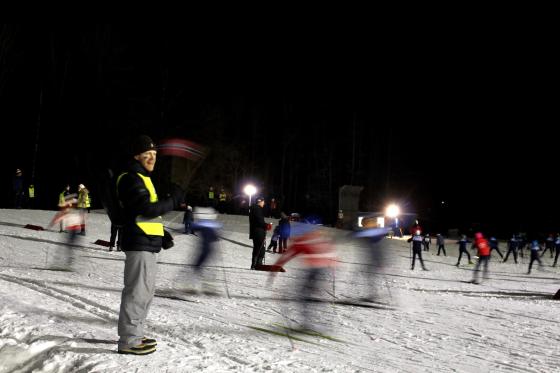 Mann st&aring;r &aring; veiver med norskt flagg foran barn som g&aring;r p&aring; langrenn.
