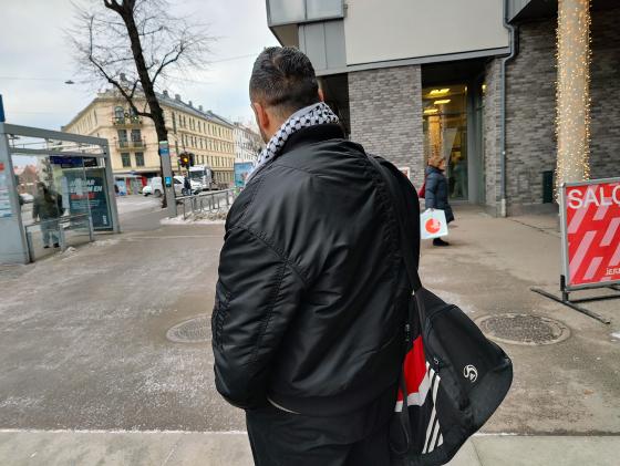 Mann med ryggen til. Sort jakke. Sort bag. Gr&aring;tt rundt.