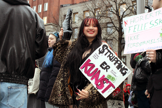 Stripper Alma Adore l&oslash;fter en strippesko i v&aelig;ret med den ene h&aring;nda og holder en plakat der det st&aring;r "Stripping er Kunst" i den andre. Hun smiler til kamera