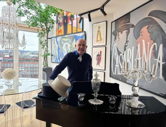 En mann bak et piano med malerier, et glassbord og vindu med utsikt ut mot Oslo bak han.