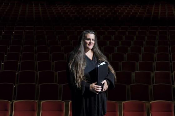 Dame står forran en teatersal, scenelyset lyser opp ansiktet hennes