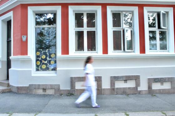 Dame i hvit sykepleieruniform g&aring;r foran en r&oslash;d bygning. Jobber i hjemmesykepleien.