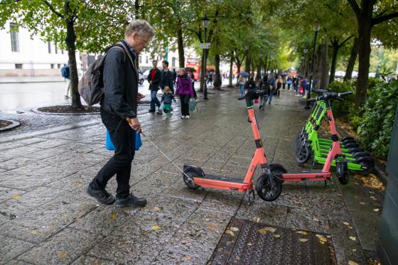 En mann med hvit stokk i høyre hånd går på et bredt fortau. Han møter på en elsparkesykkel som står parkert mer i midten, sammenlignet med resten som står parkert fint til siden. Stokken treffer elsparkesykkelen.