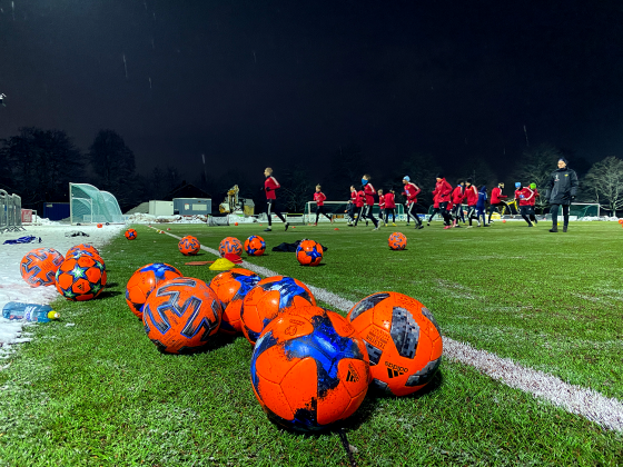 På fotballbane med spillere som trener i bakgrunn. Ser fotballer og en drikkeflaske.