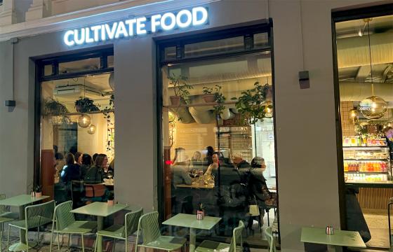 Utsiden av den veganske resturanten "Cultivate Food". Navnet i stor hvir lysende tekst over glassvinder meg folk som sitter inne til syne gjennom vinduet.