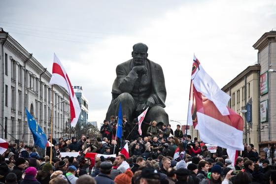 Mange demonstranter som bærer på hviterusslands hvite og røde flagg. De er vendt med ryggen mot kameraet og ser på en stålstatue av Lenin.