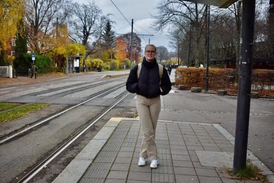 Blond kvinne med svart jakke, beige ryggsekk og mørke briller står og venter på trikken utenfor Universitetet i Oslo, avd. Blindern