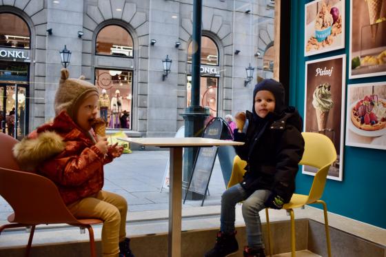 To små barn i parkdresser og luer sitter inne på en iskrembutikk og spiser is en kald novemberdag. Begge spiser en kule sjokoladeis i kjeks. Gjennom vinduet utenfor kan man se travle mennesker på Karl Johan. 