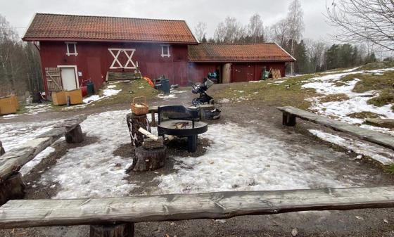 Rødt gårdsbruk om vinteren. Tre benker i front av bildet til å sitte på, med et bål med røyk i midten