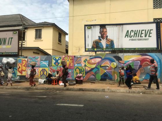 Bildet viser et fortau i Ghana. Det går flere personer på fortauet, og bak dem er en vegg dekket i fargerik graffiti. 