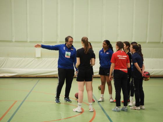 Håndballtrener peker på noe utenfor bildet og jentene står samlet i en gruppe rundt han.