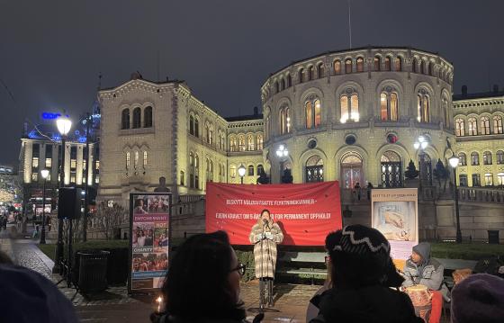 Bildet viser Khansa Ali som står foran Stortinget og holder en appell. Bak henne henger det et rødt banner hvor det står "Beskytt voldsutsatte flyktningkvinner - fjern kravet om 5 års botid for permanent opphold".