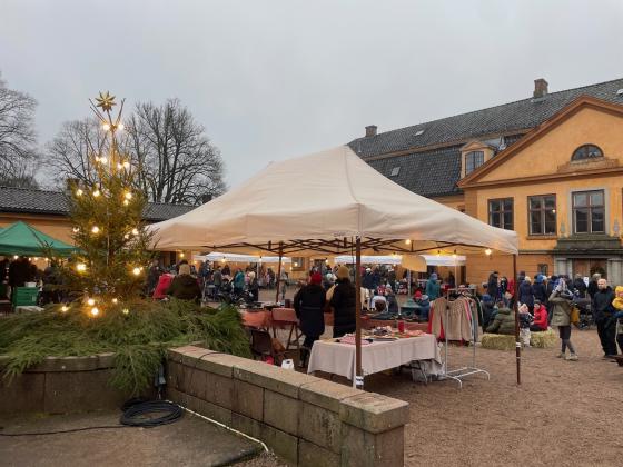 Bildet viser julemarkedet på Bogstad Gård. Man kan se et juletre med lys, et hvitt telt som holder regnet unna noen som selger produkter, og mange folk i bakgrunnen. 