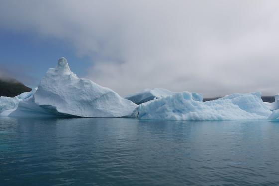 Bildet viser en liten isbre på Grønland, liggende på et stille turkist hav, over isbreen er det mørke skyer