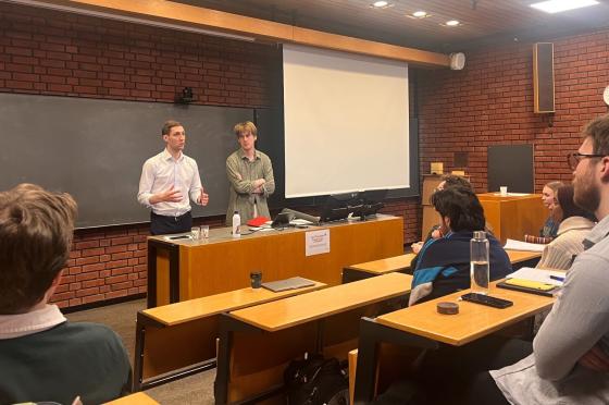 En mann i hvit skjorte står foran i et auditorium sammen men en gutt i grønn skjorte. Studenter følger med fra salen. 