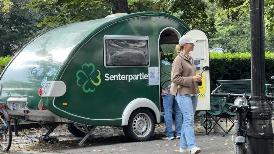 Bilde av en blond kvinne i caps, beige jakke og lyseblå jeans foran senterpartiet sin grønne campingvogn-valgbod på Karl Johan, som ser til siden