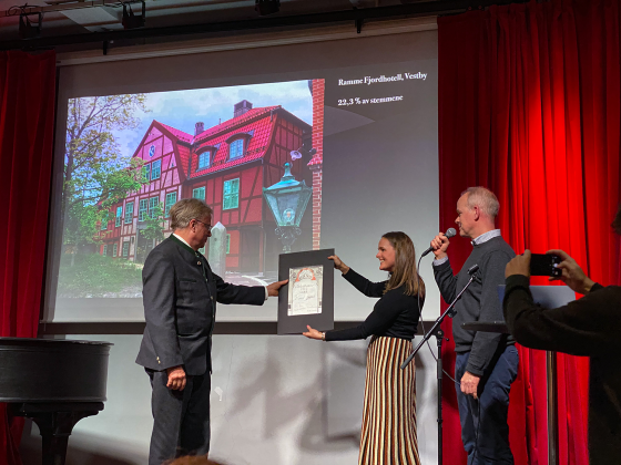 Bilde av tre folk som står på en scene. Mannen får en pris (hedersprisen). I bakgrunnen er det en powerpoint med bilde av Ramme Fjorhotell som ligger i Vestby.