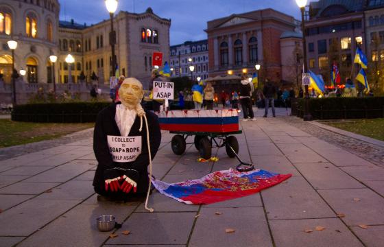 En dukke som forestiller Putin står på kne foran en metallskål. I bakgrunnen står en kiste på hjul malt etter det russiske flagget. 