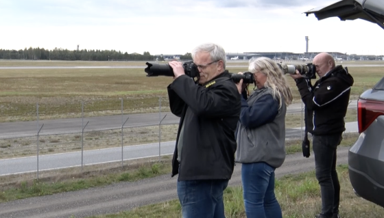 Tre flyspottere med kameraet mot ansiktet i høyre hjørnet av bildet. Rullebane på Gardermoen i bakgrunnen