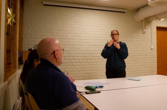 En dame med briller står foran et bord med hånden foran munnen. Munnen er formet som en O. Foran henne sitter det to mennesker med ryggen til kamera.