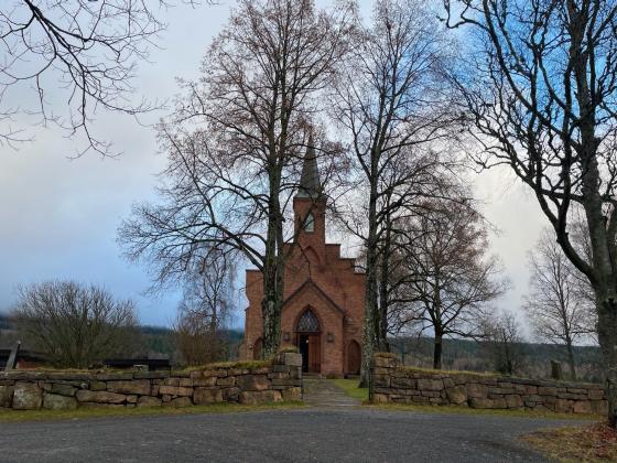 Bildet viser Sørkedalen kirke i Oslo. Kirken er laget av brune murstein, og har et høyt kirkespir. Den står litt tildekket av nakne løvtrær. 