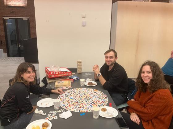 Bord med tre studenter rundt, på bordet ligger noen brettspill og papptallerkner med snacks