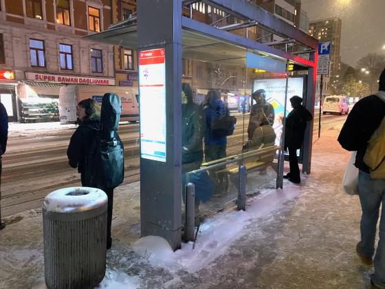 Bildet viser flere mennesker som venter på bussen på Tøyengata buss-stopp, vendt mot Super Bawan Dagligvare. Det er litt mørkt ute, og snøer faller ned. Det er snø på bakken. Personene står med ryggen til fotograf.