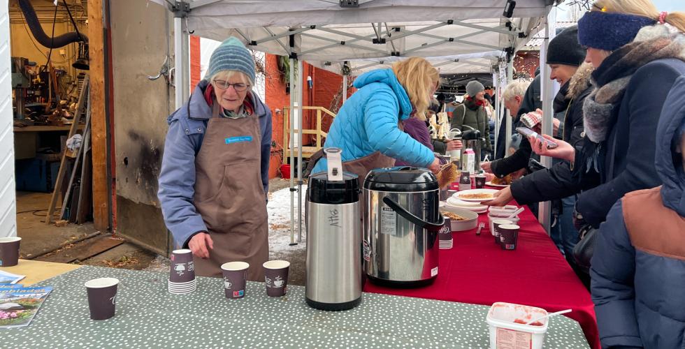 To kvinner fra venneforeningen botanisk hages venner som står bak et bord. den bakerste gjør klar en vaffel. kvinnen nærmest kamera er på vei å fylle på kaffe. utenfor bode er det folkerikt.