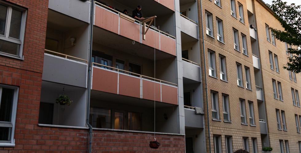 En blokk med en balkong med en hjemmelaget kran, som heiser ned en kurv med pizza esker. Det står en menneske samling og venter på gata.