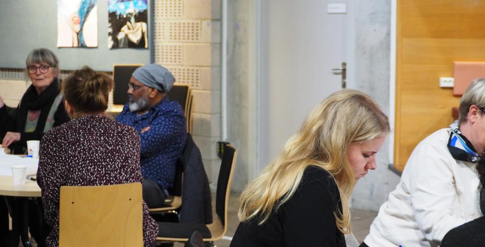 Fem mennesker som er avbildet. Tre sitter ved ett bord, og de to andre setter ved et annet. 