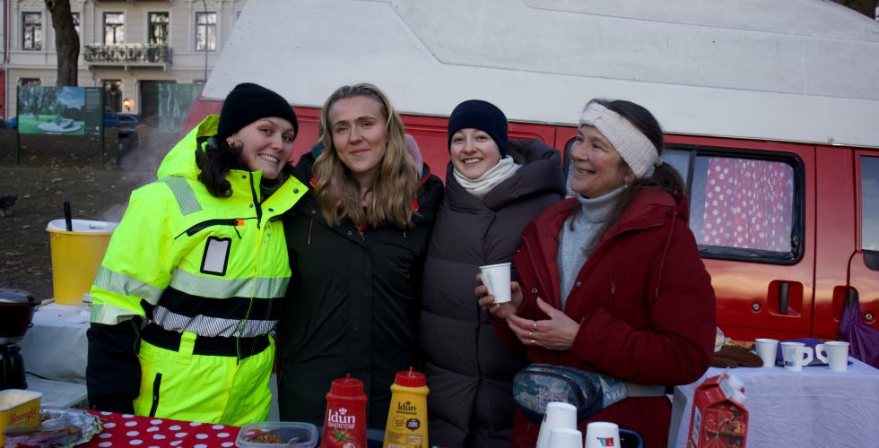 fire damer står og smiler bak kaffebordet. I bakgrunnen er en rød og hvit van 