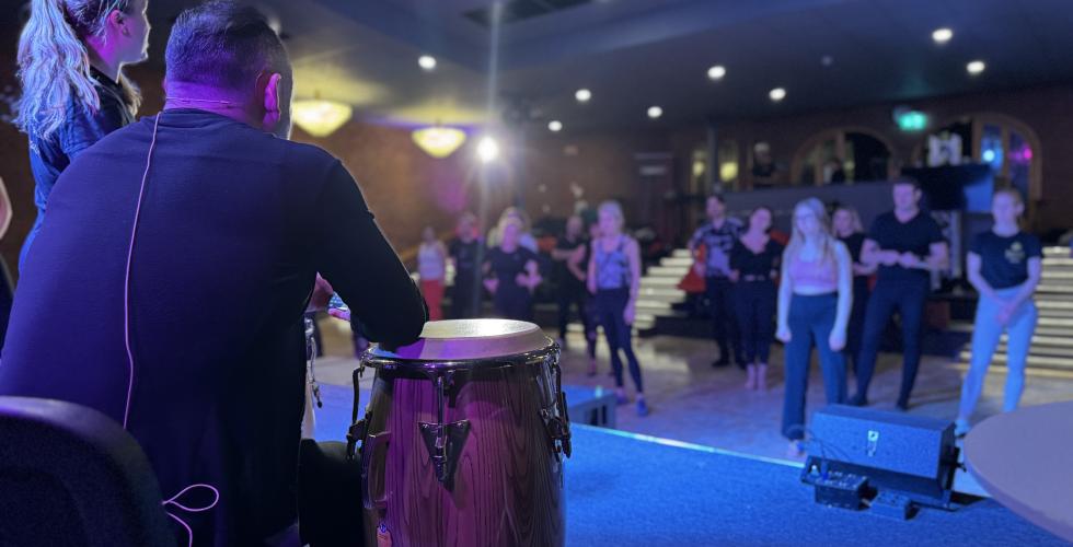 Bilde tatt fra scenen. I forgrunn sitter en instruktør og spiller trommer, mens en annen viser dans. I bakgrunnen står en mengde elever som er på nybegynnerkurs i salsa. 