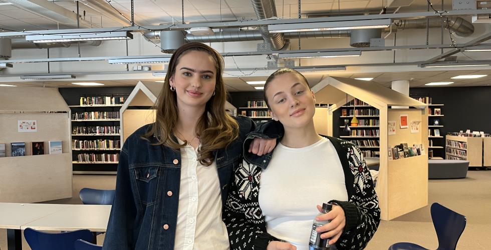 To unge jenter står ved siden av hverandre på biblioteket og smiler