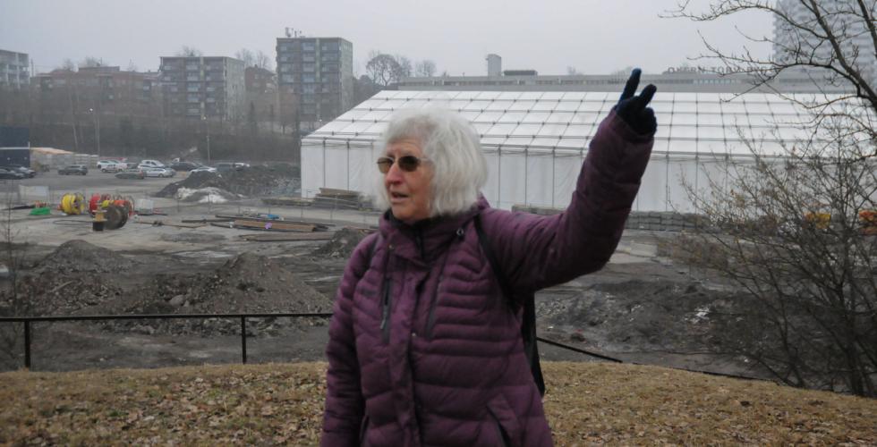 En dame med lilla boblejakke forteller om byggeplassen man ser bak henne