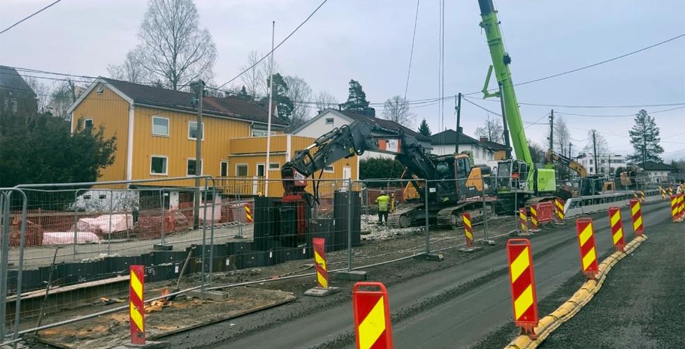 En høy grønn maskin til høyre. Foran denne maskinen står det en gul og sort gravemaskin. Rundt maskinene er det et langt gjerde av metall. I bakgrunnen ser man et gult hus. Foran maskinene er det en smal grusvei.