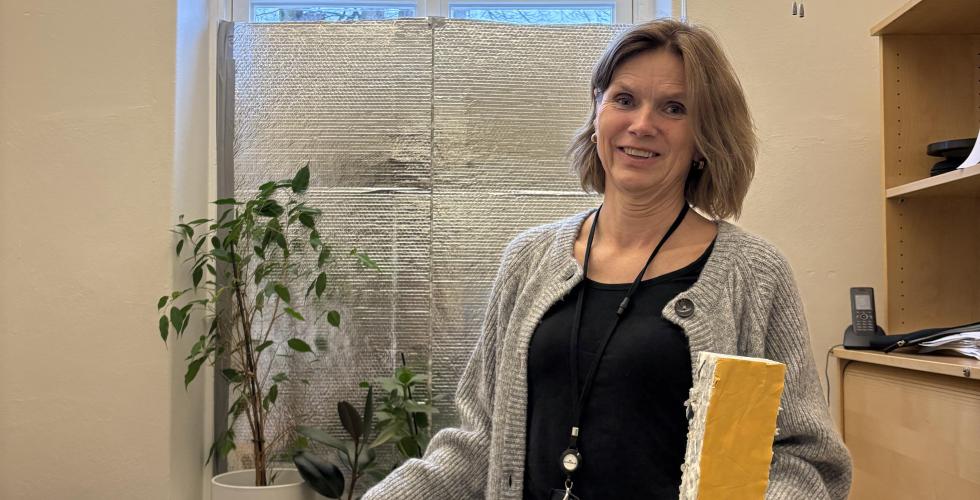 Randi Hjertaker viser frem isolasjonsplater som hun har brukt til å polstre igjen vinduet. Hun står foran vinduet på sitt eget kontor. 