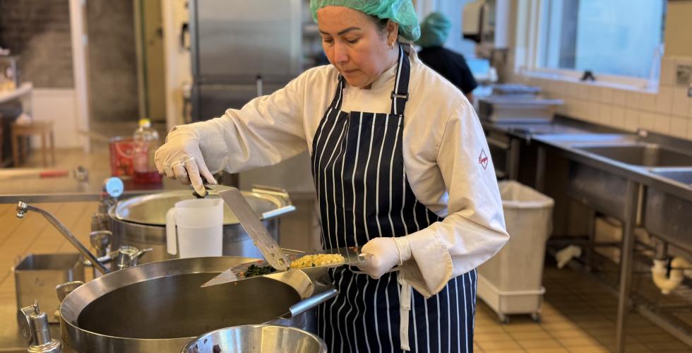 Andrea står på et kjøkkenet ovenfor en kjele og skyver grønnsaker fra en fjøl over i kjelen. Hun har på forkle og hårnett og konsentrerer seg på matlagingen. 