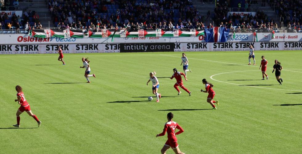 To fotballlag spiller mot hverandre på en kunstgressbane. Det palestinske laget har rød drakter. Vålerenga sitt lag har hvite drakter. Det er tilskuere på tribunen.  