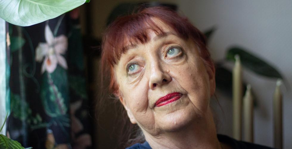 Portrett av en eldre dame med rødt oppsatt hår og pannelugg. Hun har blikket rettet mot et vindu. I bakgrunnen ser man en plante, noen lys og gardiner