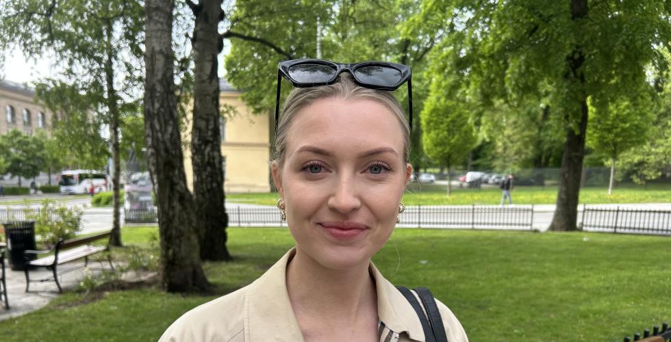Ung dame som smiler med grønn natur i bakgrunnen imens hun venter på bussen.
