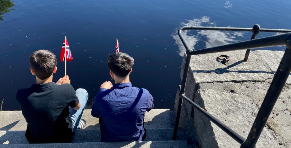 To personer sitter i trapp nede ved vannet med hvert sitt norske flagg