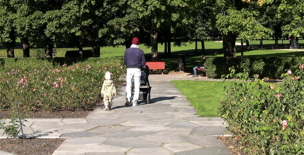 Far triller barnevogn nedover en sti i en park, et lite barn går ved siden av