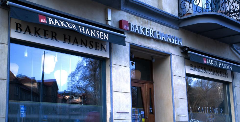 Bildet viser fasaden til en Baker Hansen-kafé med sort markise, røde «1861»-detaljer og store utstillingsvinduer som speiler trær og bygninger på andre siden av gaten. Den aktuelle Baker Hansen-kafeen på bildet er nå nedlagt.